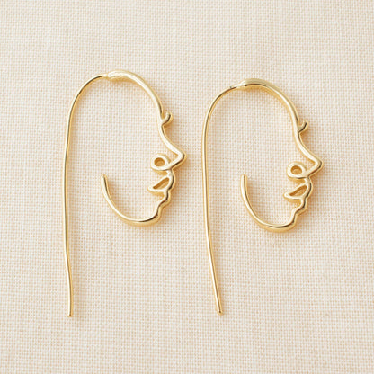 Gold face earrings with a neutral background. 